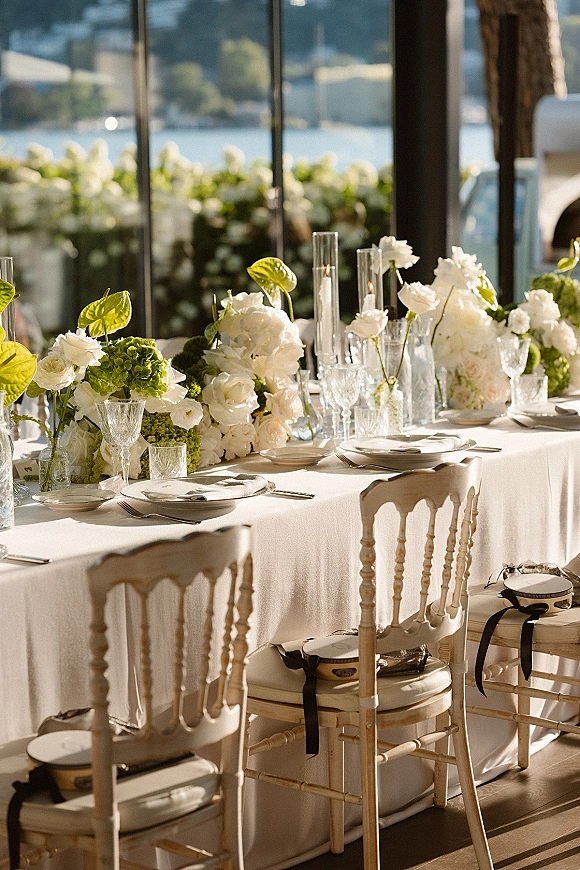 Reception tablescape with wedding head table decor, white and green centerpieces, tall cylinder candles, and tambourines by sunlit windows overlooking water