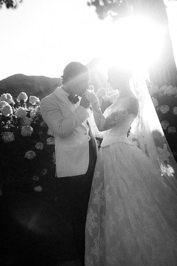 Wedding couple portrait of groom kissing bride’s hand as she wears lace sleeves and a long veil, backlit in a garden with mountains and sun flare