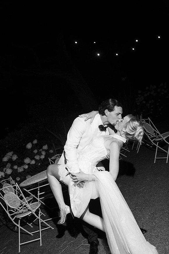 Wedding kiss in a dramatic dip, groom in a white tuxedo and bride in a strapless dress beneath string lights in a garden at night