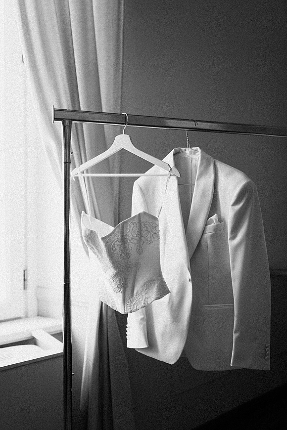 Wedding attire flatlay of a white tuxedo jacket and lace corset on hangers by a window, with pocket square on a clothing rack