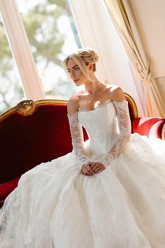Bridal portrait of a bride in an off the shoulder wedding dress with lace sleeves, seated on a red velvet settee by a window in natural light