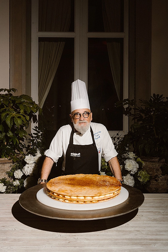 Wedding cake on a dessert stand as a chef in a hat and apron presents it by a window with curtains and potted greenery indoors