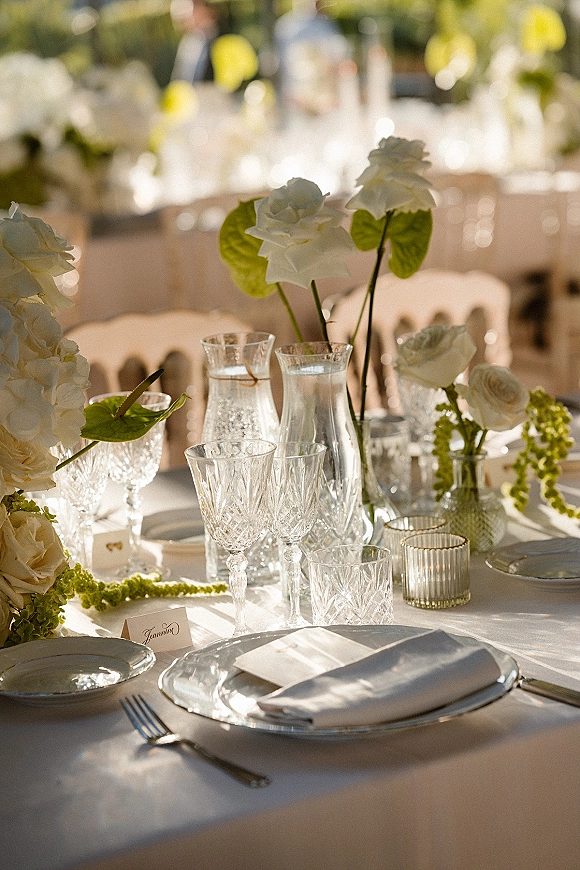 Reception tablescape with wedding table setting details, white floral centerpieces, crystal stemware, and votive candles in sunlight outdoors