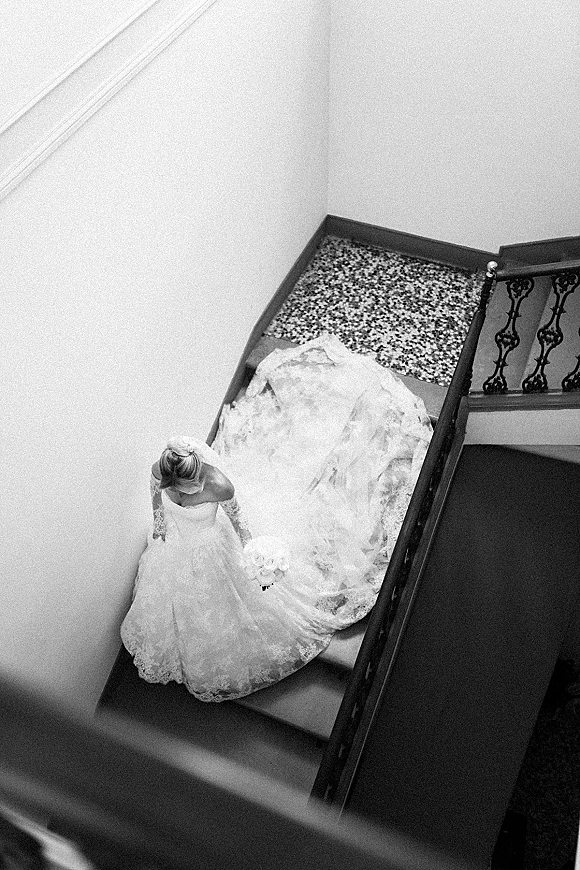 Bridal portrait of a bride on staircase holding a bouquet, her lace long-sleeve gown with dramatic train spread on terrazzo floor