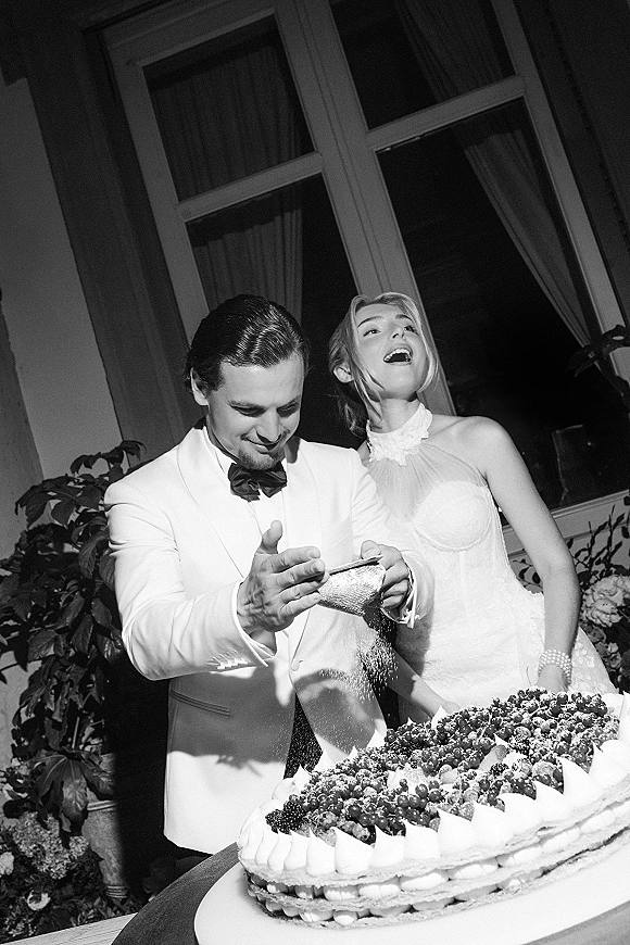 Wedding cake moment as bride laughs while groom in white tux and black bow tie sprinkles powdered sugar over berry-topped cake by window curtains