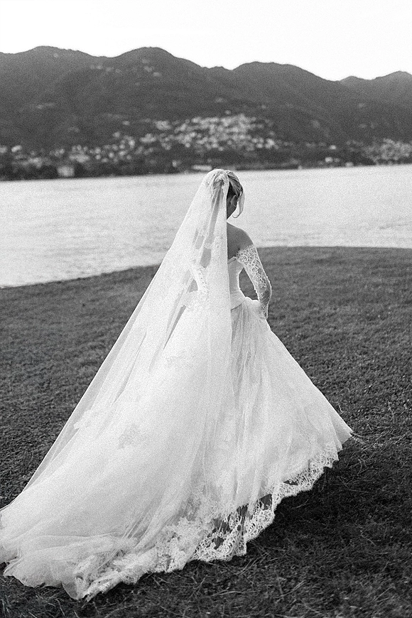 Bridal portrait of a bride from behind holding her lace train, long cathedral veil flowing by a lakeside with mountains in view