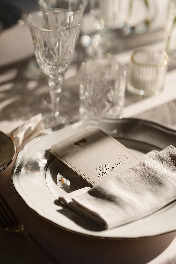 Reception tablescape with wedding place setting featuring a menu card on the plate, linen napkin and crystal glassware on tabletop