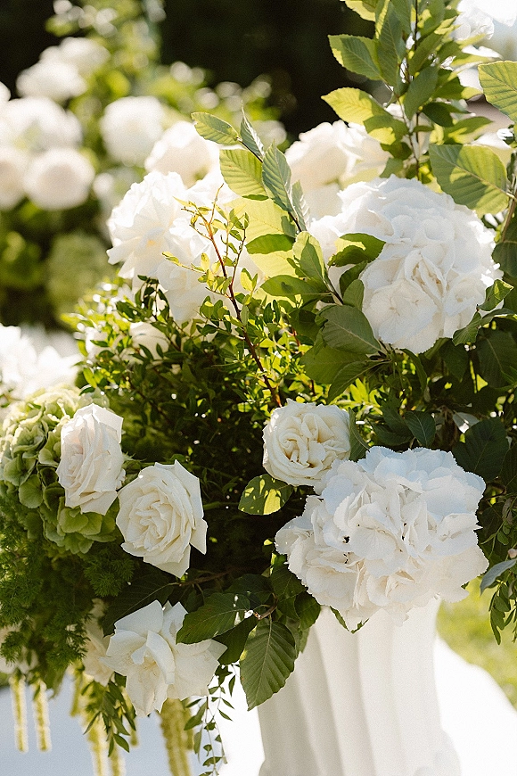 Wedding floral arrangement of white wedding flowers with roses, hydrangeas, and greenery on a white pedestal in sunlit garden greenery