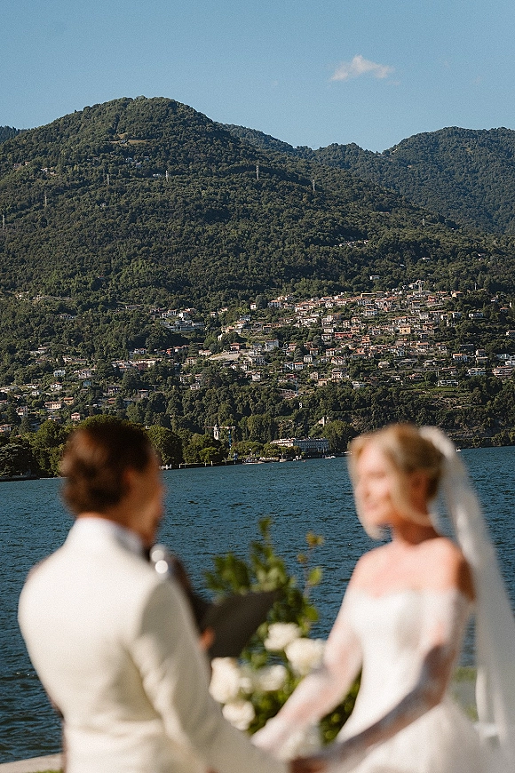 Wedding ceremony moment at an outdoor lakeside wedding with bride and groom holding hands, veil flowing, lake and mountains behind