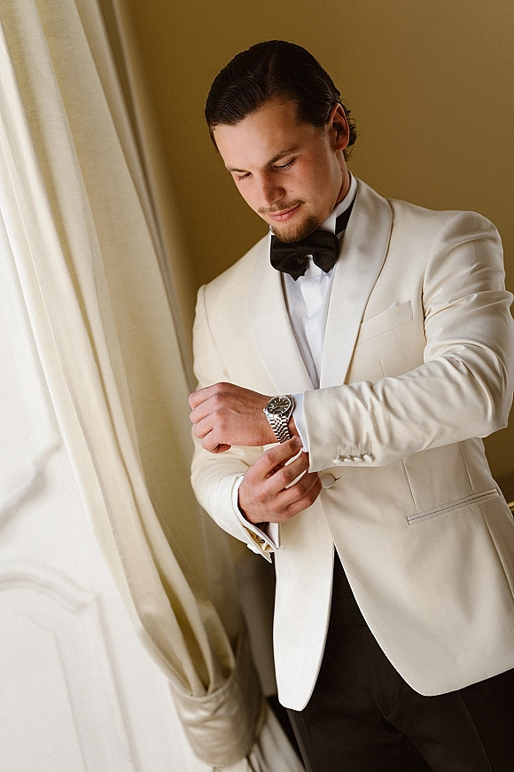 Groom portrait in an ivory tuxedo jacket and black bow tie, adjusting cufflinks by a window with sheer curtains and soft light