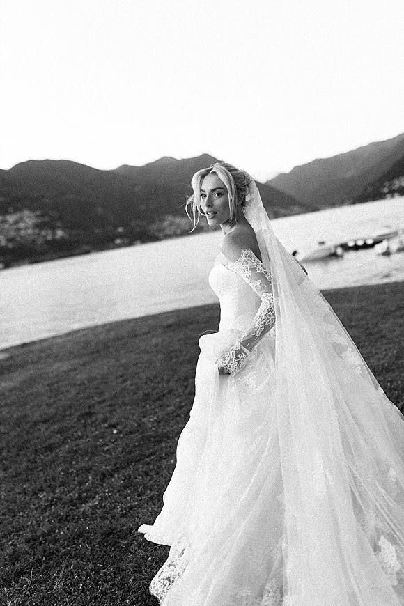 Bridal portrait of a bride in a strapless lace wedding dress with cathedral veil, looking over her shoulder by a mountain lake shoreline