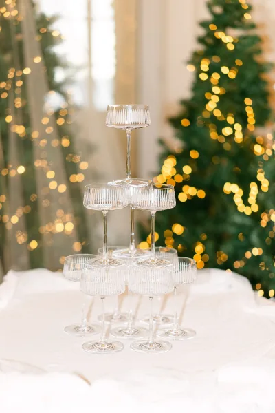 Champagne tower wedding display of stacked coupe glasses on a white tablecloth, set by Christmas trees with twinkle string lights and window glow