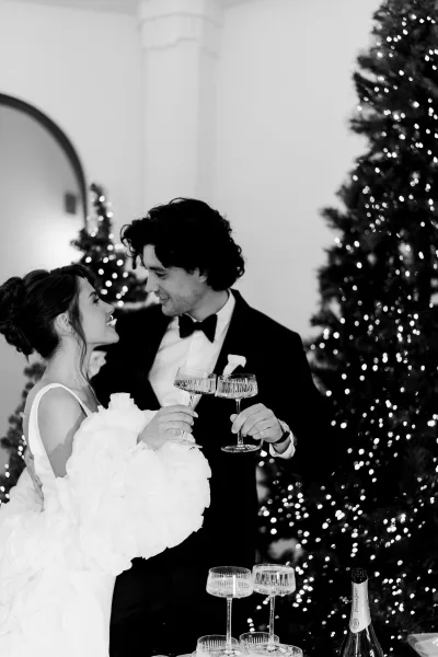 Wedding toast as newlyweds toasting with champagne coupe glasses, bride in ruffled gown and groom in tuxedo by lit Christmas trees indoors