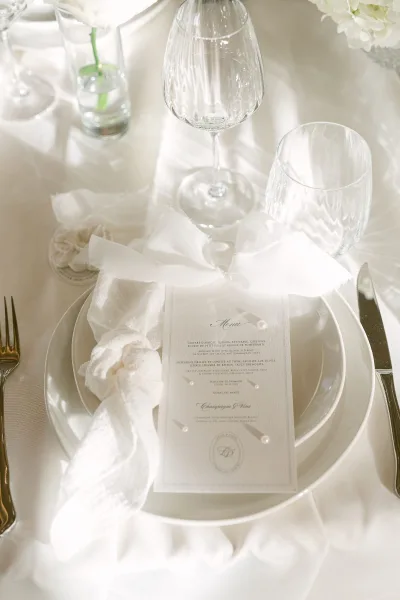 Wedding tablescape with layered white plates and a menu card with pearl accent, gold flatware, crystal glasses, and bud vase in natural light