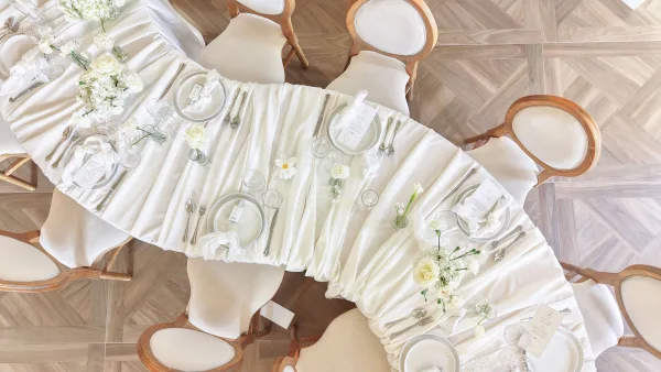 Reception tablescape with white wedding table setting, menus and place cards, bud vases of white florals on a parquet floor backdrop