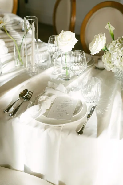 Reception tablescape with wedding place setting, white taper candles in glass holders, menu card, and white rose and hydrangea bud vases indoors