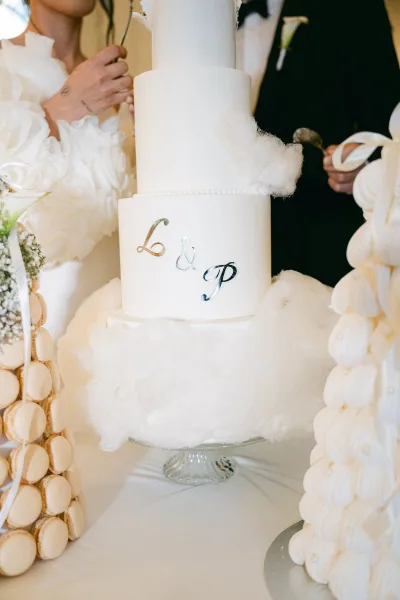 Wedding cake with white tiered wedding cake layers and monogram letters on a glass stand, surrounded by cotton candy and macaron towers at an indoor dessert table