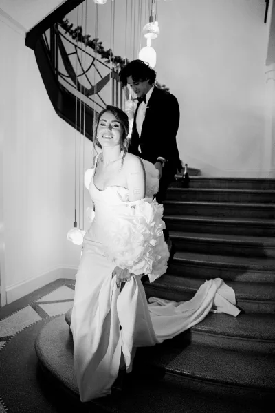 Couple portrait of bride on staircase holding a calla lily bouquet as groom stands behind her, bridal train draped on stairs under pendant lights