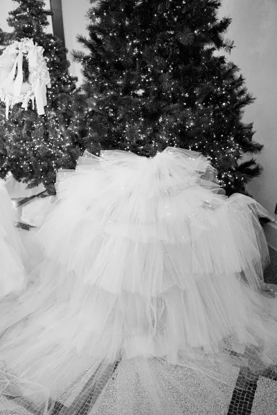 Wedding dress with tulle wedding dress ruffle skirt spread on a tiled floor beside Christmas trees with string lights and wreath ribbon accents