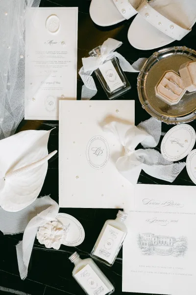 Invitation suite wedding invitation flat lay with monogram crest, wax seal, pearls, sheer ribbon, rings, shoes, and perfume on a dark tabletop