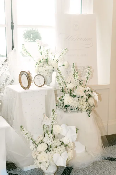 Wedding welcome sign with florals on an easel beside a pedestal of white roses and calla lilies, framed table cards, pearl tulle drape by a large window