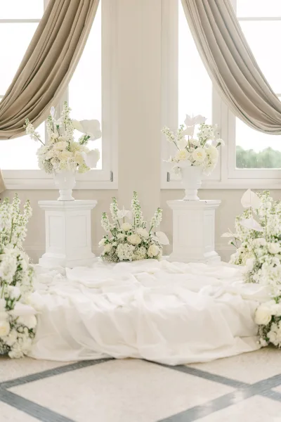 Ceremony altar decor with wedding altar flowers in white urn pedestal arrangements, fabric draping, and aisle florals before large windows