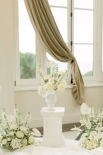 Wedding floral decor with white wedding florals on pedestal stands in urn vases, roses and calla lilies by draped windows and greenery view
