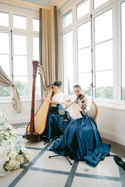Wedding musicians perform wedding ceremony music as a harp and violin duo in navy gowns by tall windows, reading from a sheet music stand