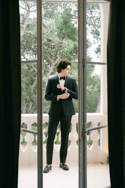 Groom portrait in a black tuxedo adjusting his wristwatch by a large window, looking away with balcony railing and trees beyond