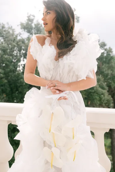 Bridal portrait of a bride in a one shoulder wedding dress with ruffled tulle, holding a pearl handle heart bag on a terrace by stone balustrade