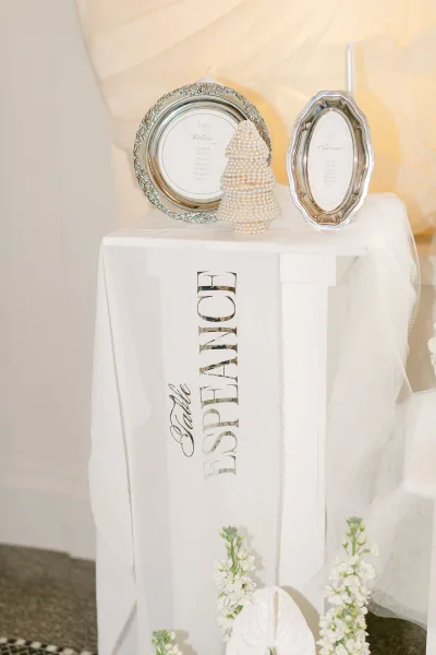 Wedding seating chart in a silver frame on a pedestal stand with calligraphy vellum, white florals, and draped fabric against a white wall
