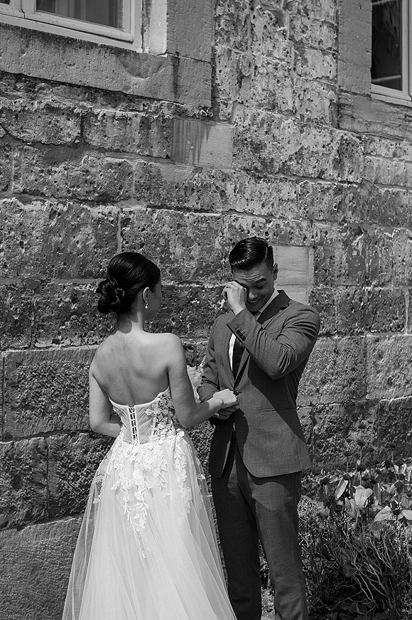 First look moment as bride in strapless lace gown holds hands with groom in suit wiping tears by a stone wall and garden plants