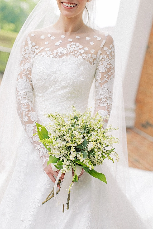 Bridal portrait of a smiling bride in a lace sleeve wedding dress, veil over shoulders, holding lily of the valley bouquet on a wooden deck