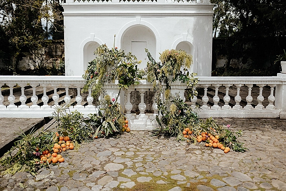 Wedding ceremony backdrop with a greenery ceremony backdrop of cascading foliage, hanging amaranthus, and citrus accents against white stucco arches