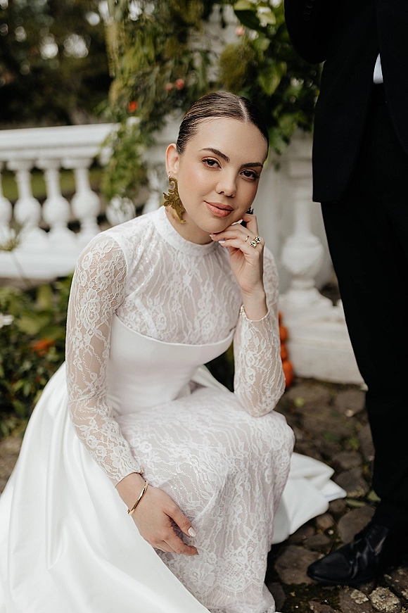 Bridal portrait in a lace wedding dress, long-sleeve high-neck lace gown with veil and gold statement earrings by garden balustrade