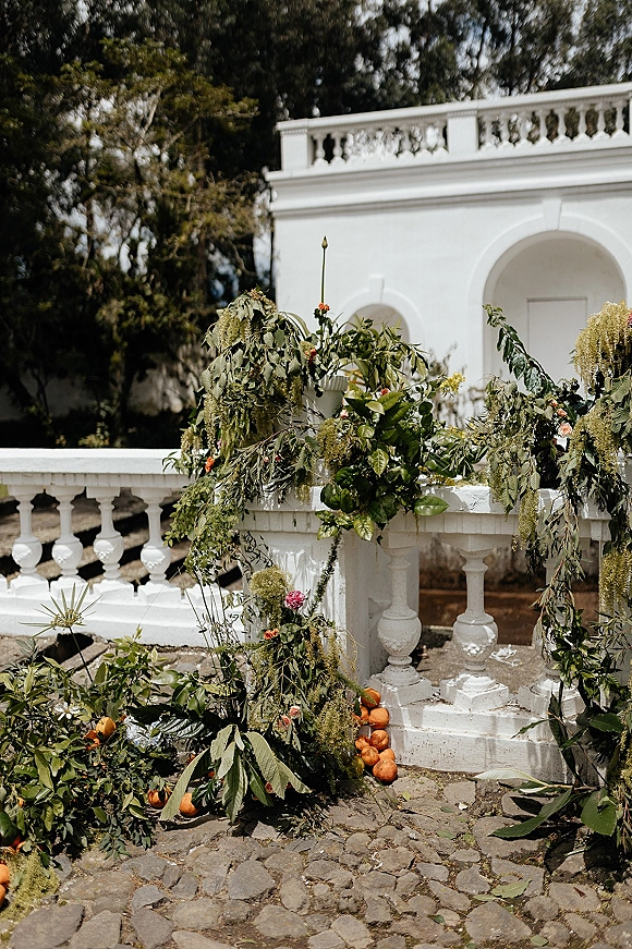 Wedding ceremony backdrop with greenery ceremony backdrop, lush garland and asymmetrical florals with oranges on a stone terrace by an arched doorway