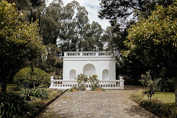 Outdoor ceremony backdrop with lush floral arrangement and greenery garland, accented by citrus fruit along a white stucco courtyard wall