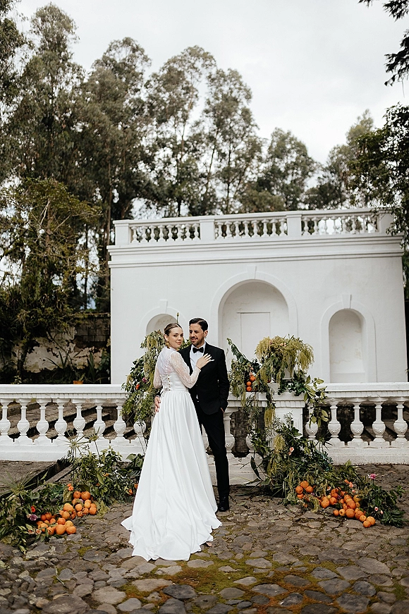 Couple portrait of bride and groom pose under a greenery garland arch with citrus accents in a cobblestone courtyard by white stucco walls