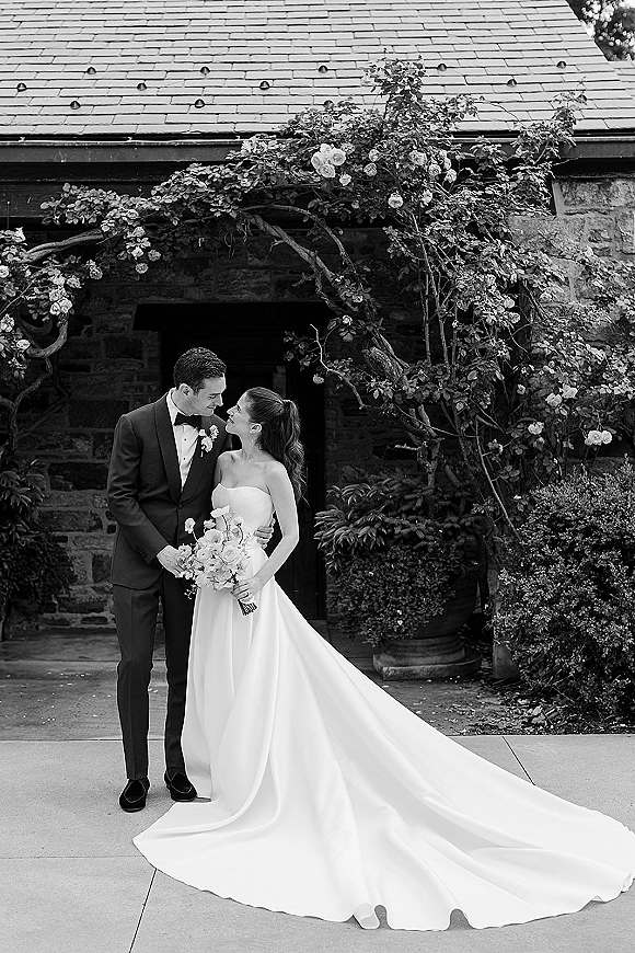Couple portrait of bride in strapless gown with long train and groom in tuxedo holding rose bouquet by stone doorway and vine