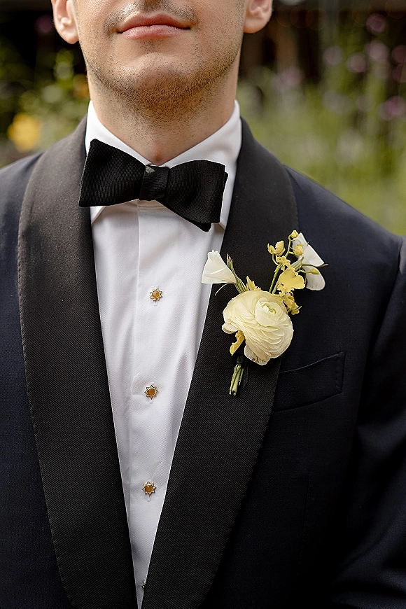 Groom tuxedo with black bow tie and white ranunculus boutonniere on a black jacket, set against soft outdoor greenery and blurred flowers