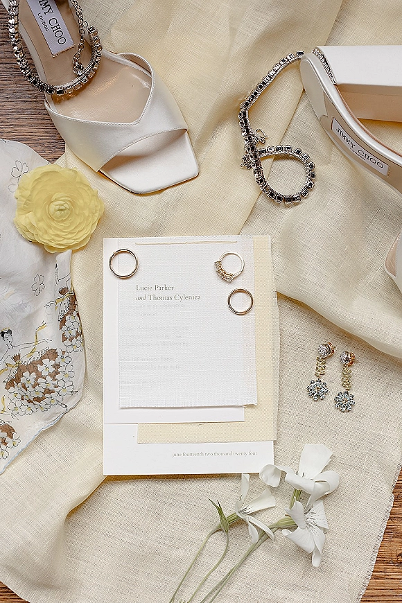 Invitation suite wedding invitation flat lay with envelope, rings, satin heels, jewelry and yellow ranunculus on a wood tabletop