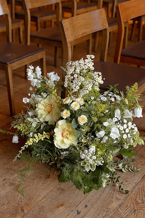 Ceremony aisle flowers in a low aisle floral arrangement with yellow and white blooms and greenery beside wooden chairs on a wood floor