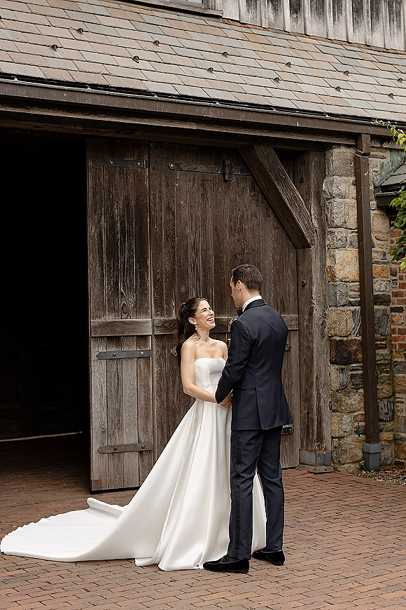 Couple portrait of bride and groom holding hands, her strapless dress train flowing as they stand by wood barn doors in a brick courtyard
