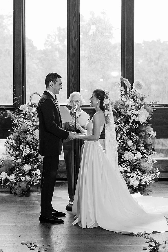 Wedding vows as bride in veil and groom in tux hold hands by a microphone, framed by window light and floral aisle petals indoors