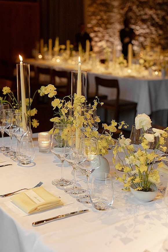 Reception tablescape with yellow wedding centerpieces, bud vases, taper and hurricane candles on white linens in a warmly lit indoor banquet space