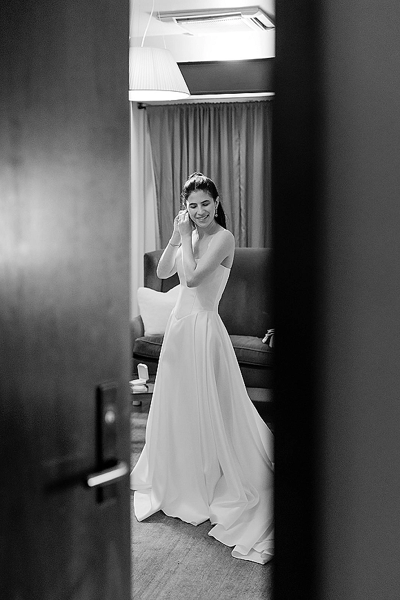 Bridal portrait of a bride in a strapless wedding dress adjusting earrings, standing by curtains in a cozy indoor room with sofa and lamp