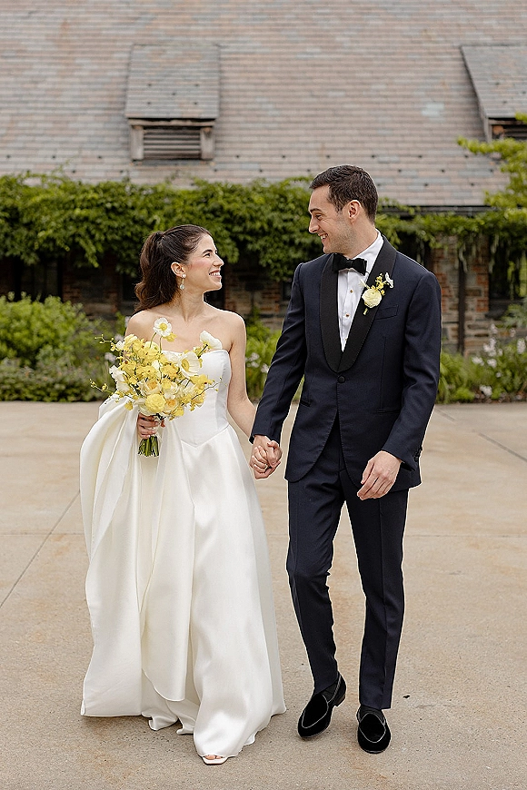 Couple portrait of bride and groom holding hands, bride in strapless gown with bouquet in a stone courtyard with greenery accents