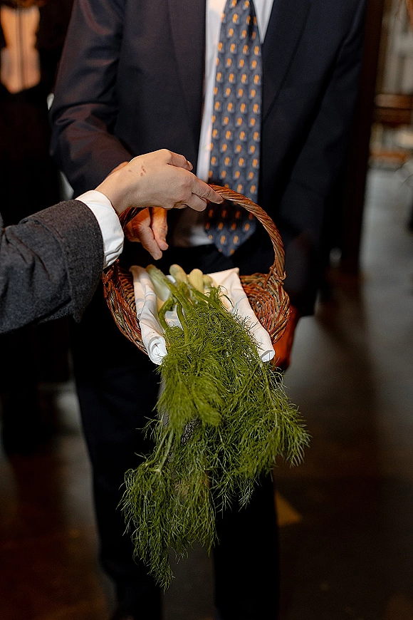 Wedding flower basket lined with white cloth and filled with greenery, held by a person in a suit jacket and patterned tie indoors