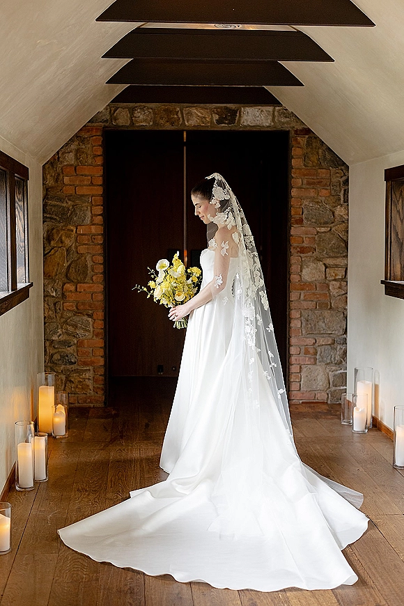 Bridal portrait of a bride holding bouquet in a strapless gown with lace veil and candlelit glass vases by a stone wall hallway