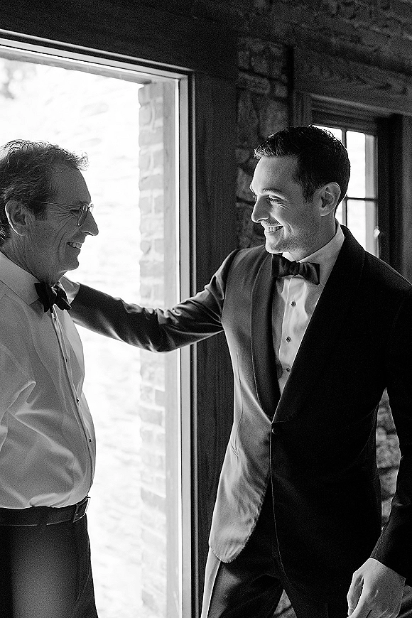 Groom with father sharing a father son wedding moment as dad adjusts cufflinks on his black tuxedo by window light against a stone wall